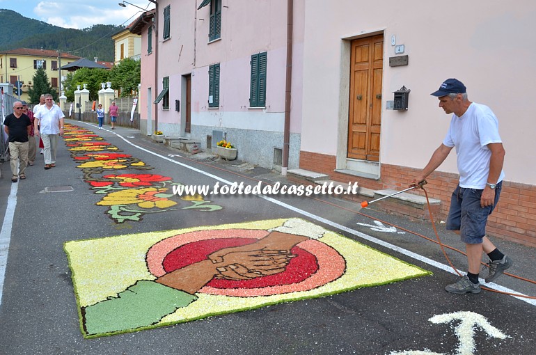 BRUGNATO (Infiorata del Corpus Domini 2015) - In Via Roma, per mantenere freschi i fiori sminuzzati, un addetto della Pro Loco spruzza acqua su un disegno di mani che si stringono in segno di pace